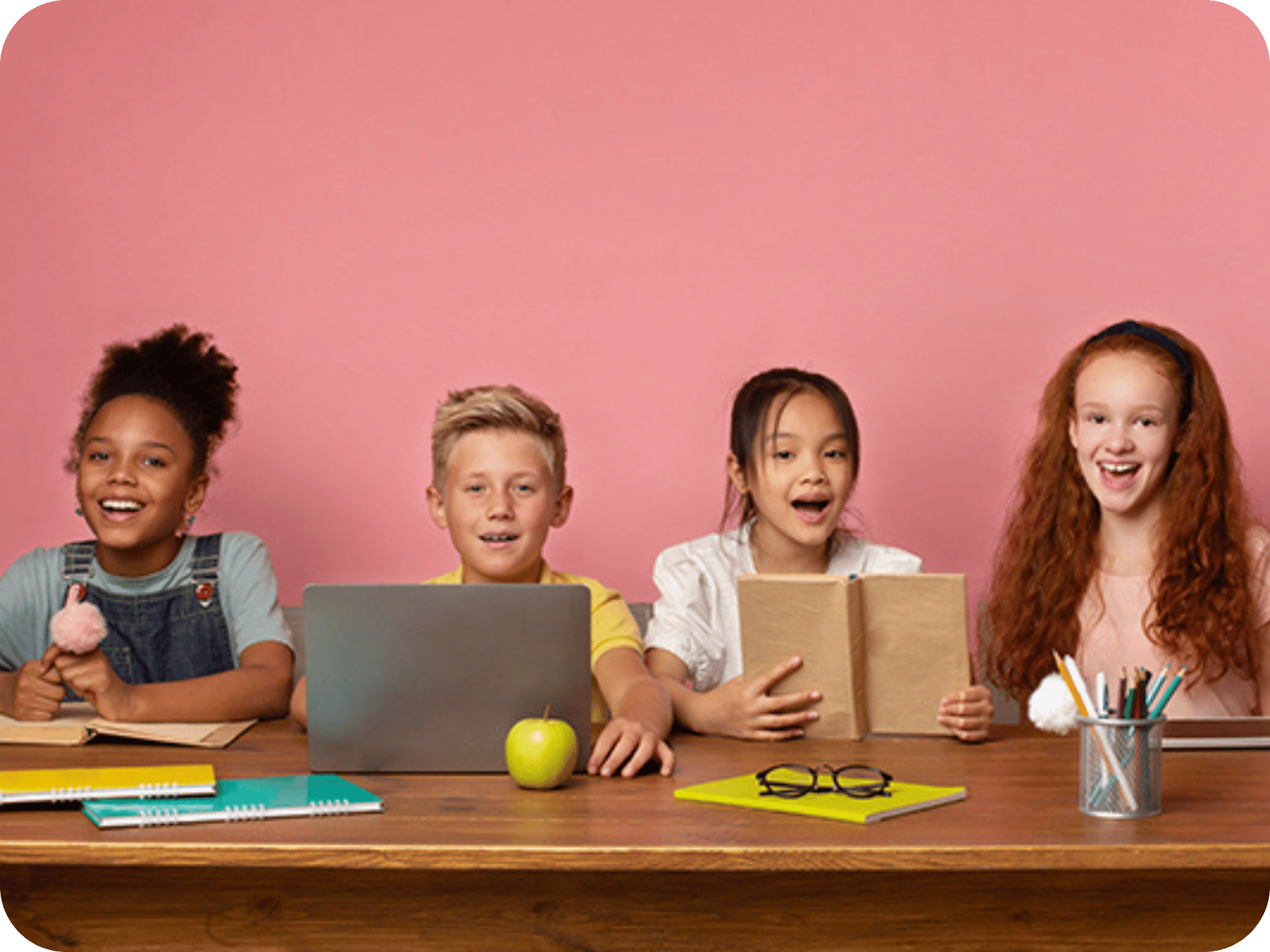Group of diverse students sitting at a desk with books and laptops, smiling and engaged against a pink background.
