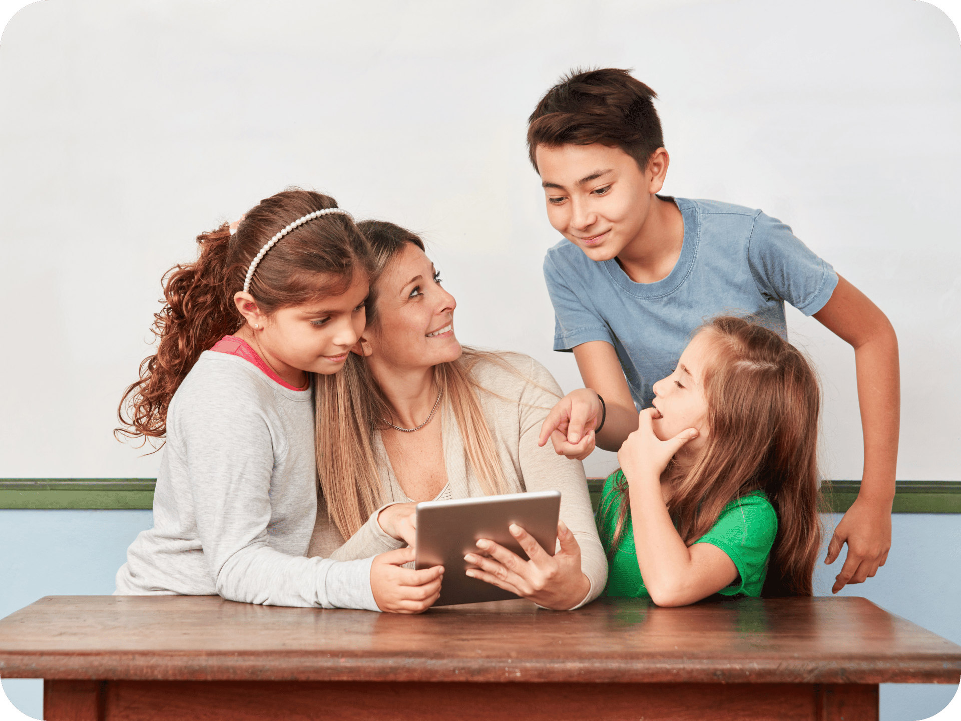 Teacher sitting at a desk with three primary-aged students, all smiling and looking at a tablet together in a classroom setting.