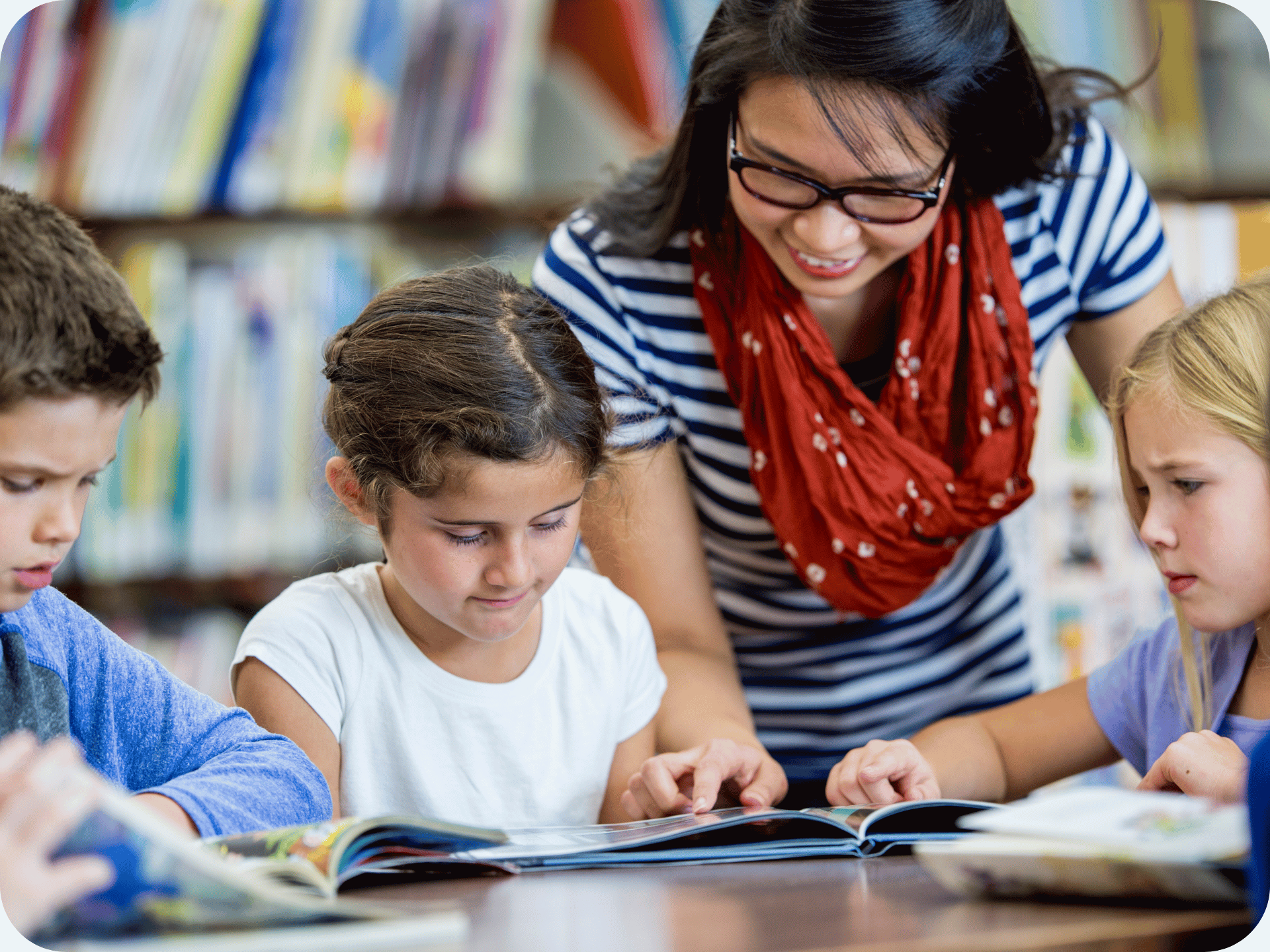 Teacher supporting a group of young students reading books together in a school library.