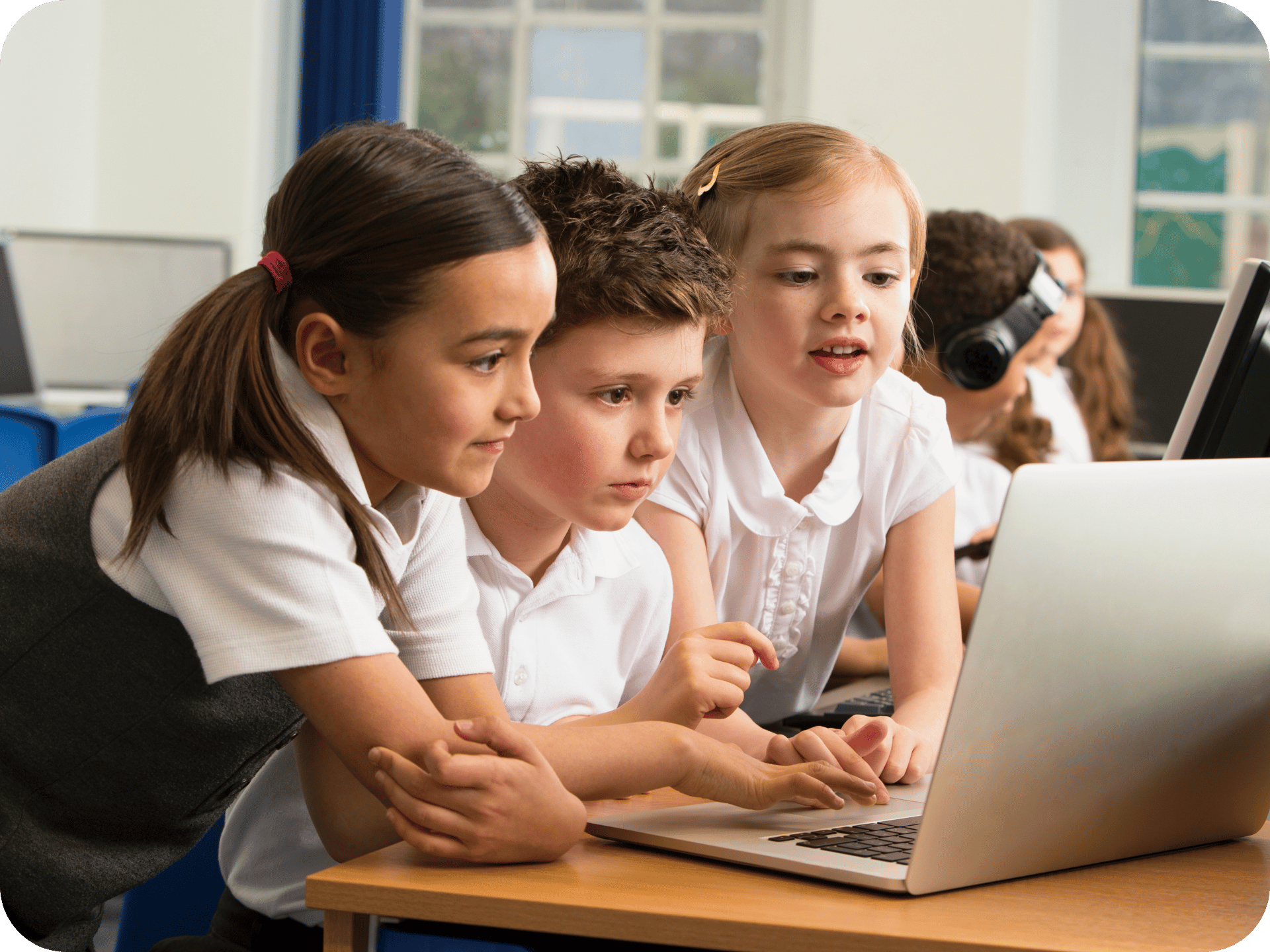 Three primary school students in uniform using a laptop together in a classroom.