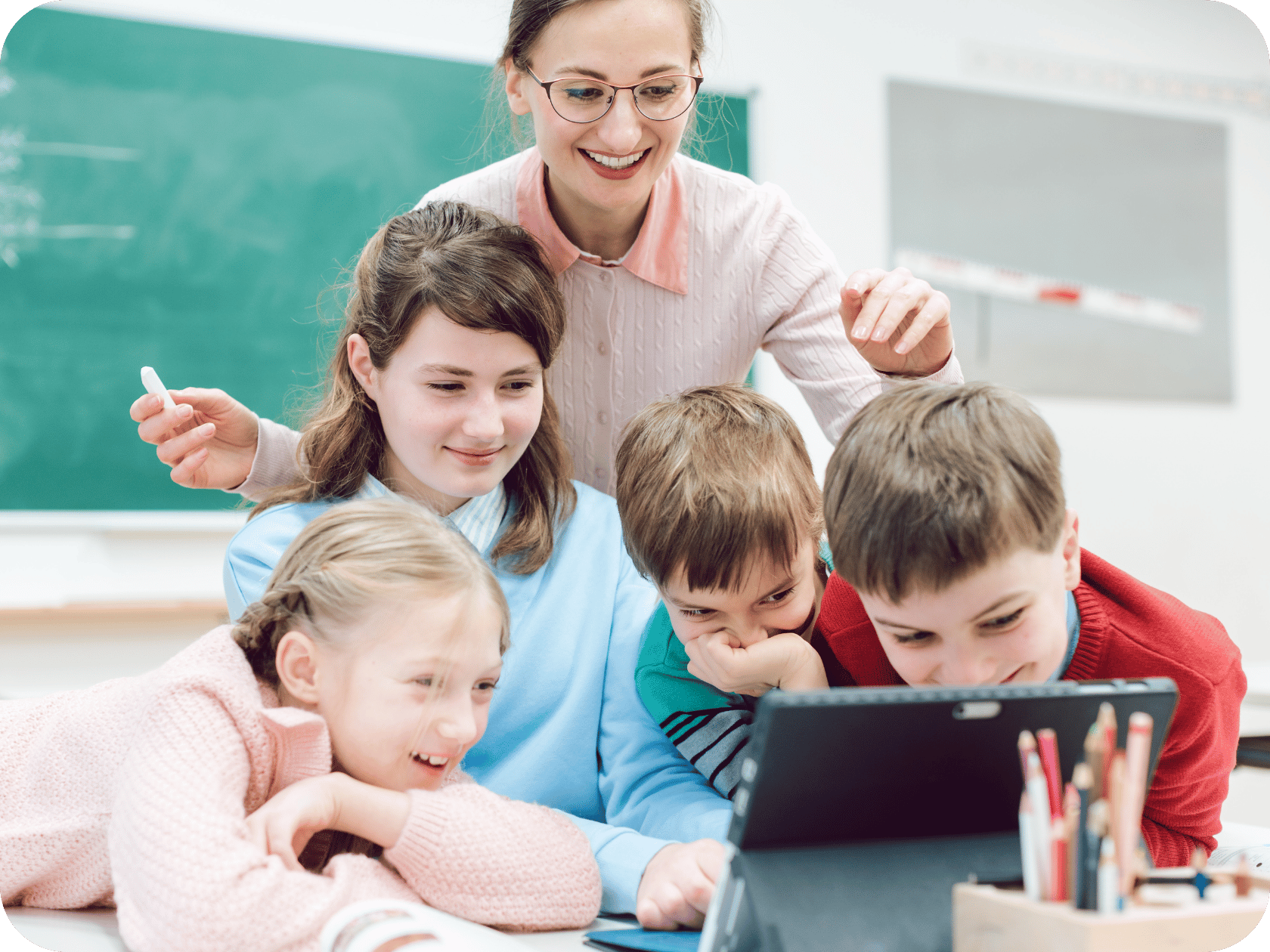 Small school teacher with a group of young students gathered around a laptop, smiling and engaged in a digital learning activity in the classroom.