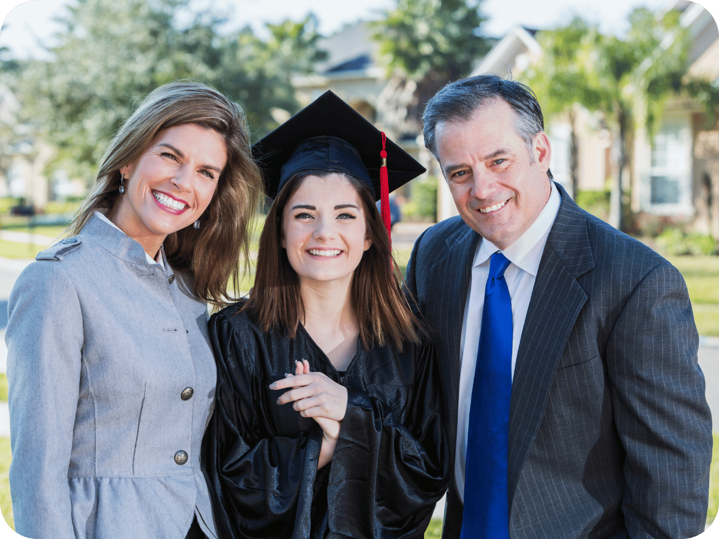 Smiling high school graduate in cap and gown standing outdoors with proud parents, symbolising student achievement and family support in education.