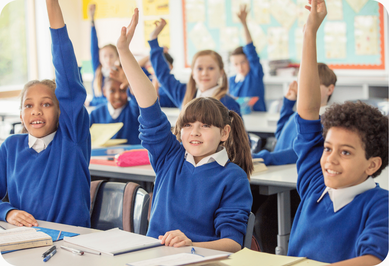 Small school students in blue uniforms raising their hands eagerly in class, seated at desks with open notebooks.